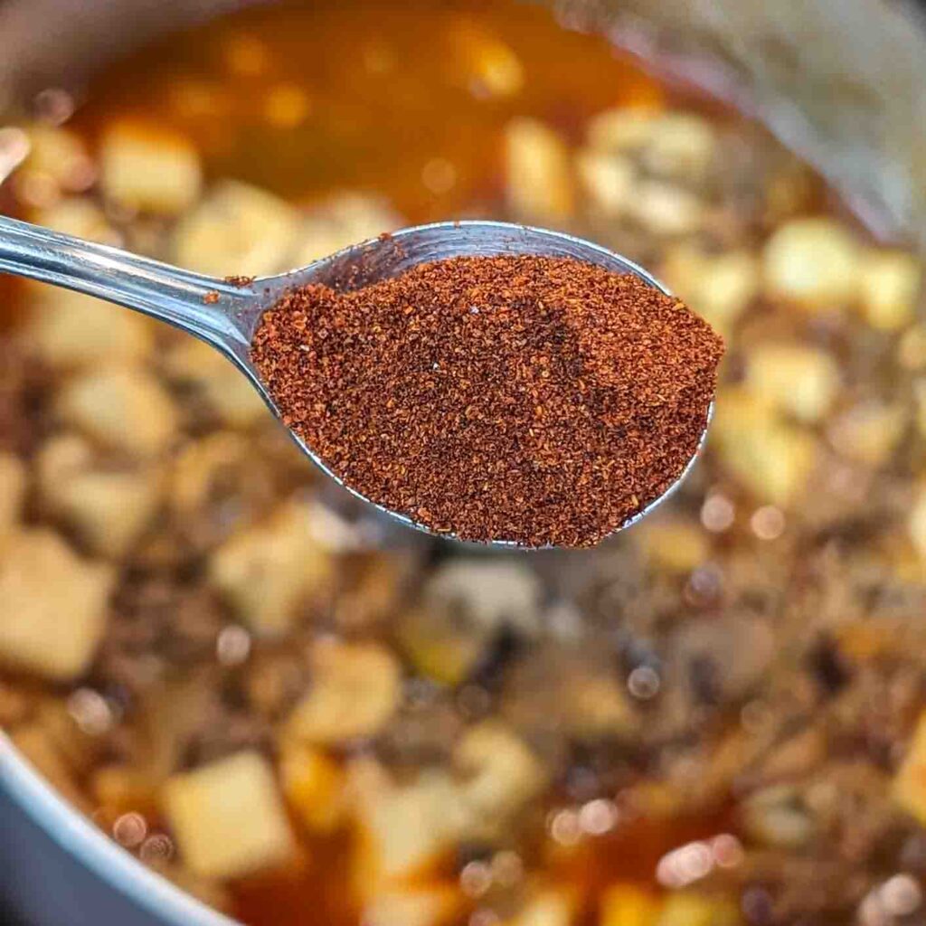 a spoonful of hot Hatch red chile powder about to be dumped into a pot of simmering Chinese Mapo Tofu